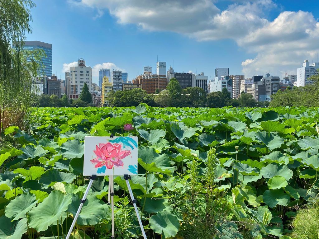 Easel with a painting of a flower before a scene of green plants and a city skyline.