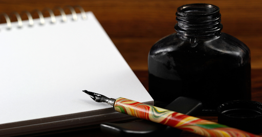 Dip pen resting on a paper pad next to a black pot of ink.